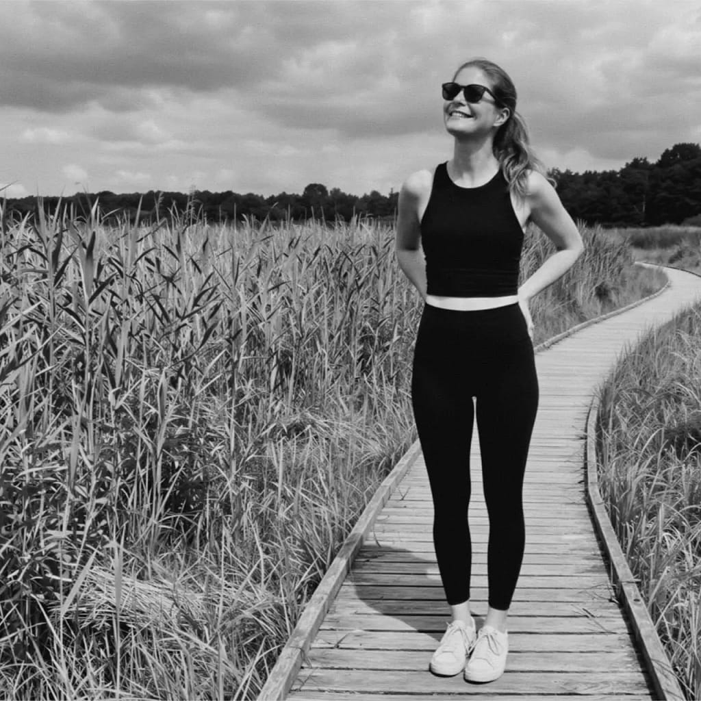 Black-and-white photo of Lady Franziska standing on a wooden boardwalk in a natural landscape, wearing sunglasses and athletic clothing.