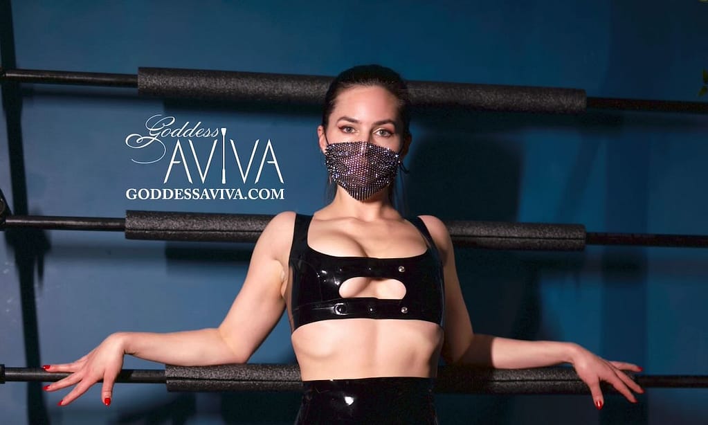 Goddess Aviva Diamond, New York City Dominatrix, poses in black latex with a jeweled face mask in a BDSM studio.
