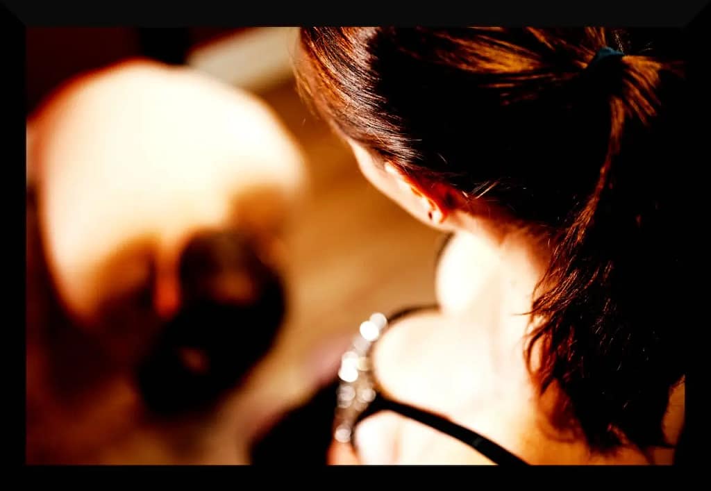 A woman with brown hair looks down at a submissive partner kneeling on the floor in a dimly lit room, symbolizing dominance and submission in a cuckolding relationship.