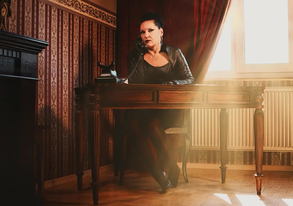 Mistress Silvia sitting behind a dark wooden desk while speaking on a vintage telephone in an elegant BDSM-inspired room with warm lighting and classic interior design.