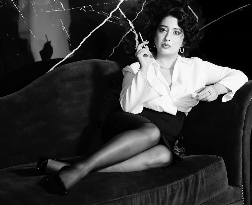 Mistress Leonor, a professional dominatrix in Barcelona, reclining on a dark velvet sofa in a white blouse, black miniskirt, sheer stockings, and high heels.