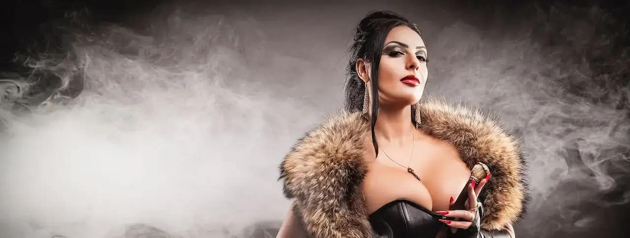 Mistress Ezada Sinn im schwarzen Korsett mit Fellstola vor rauchigem Hintergrund.