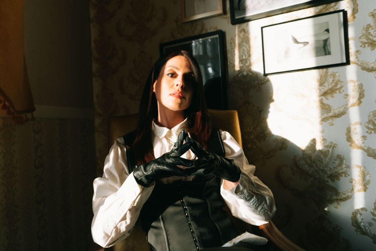 Mistress Aido Akida, a Berlin-based dominatrix, seated in dramatic light wearing a white shirt, black corset, and black leather gloves.