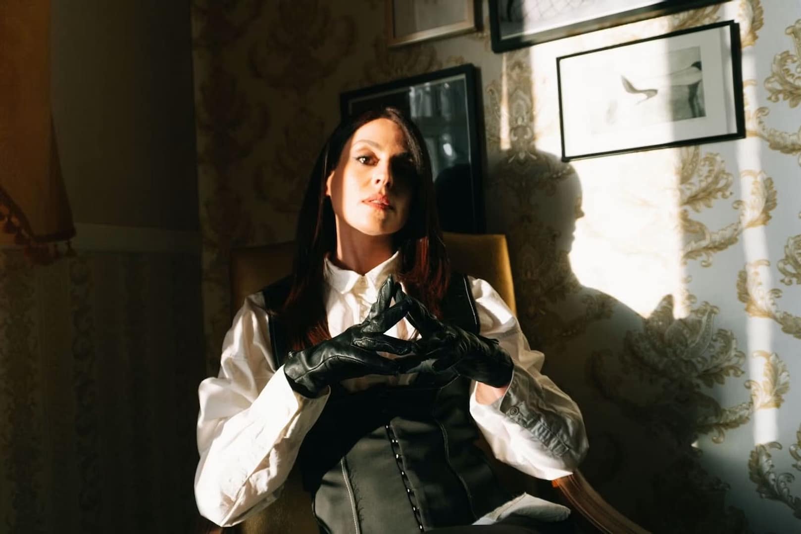 Mistress Aido Akida, Domina in Berlin, sitzt in weißer Bluse, schwarzem Korsett und Lederhandschuhen in dramatischem Licht.