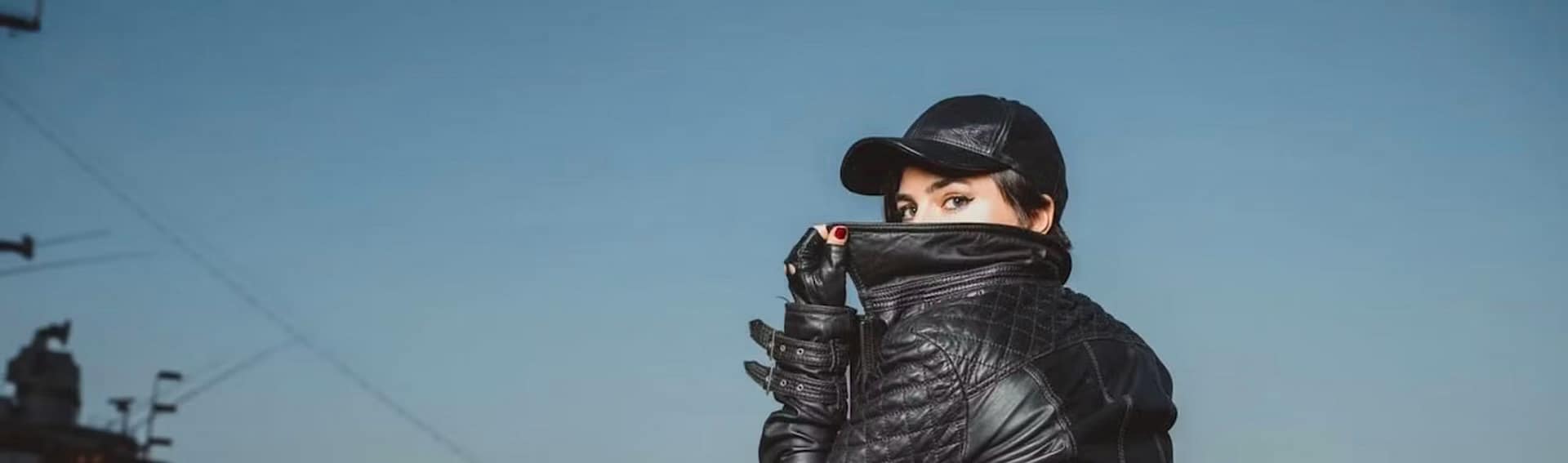 Domina Goddess Aviva trägt eine schwarze Lederjacke, Kappe und Handschuhe und blickt vor blauem Himmel direkt in die Kamera.