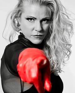 Dominatrix Lady Nastasia in black with a red latex glove, looking directly into the camera.