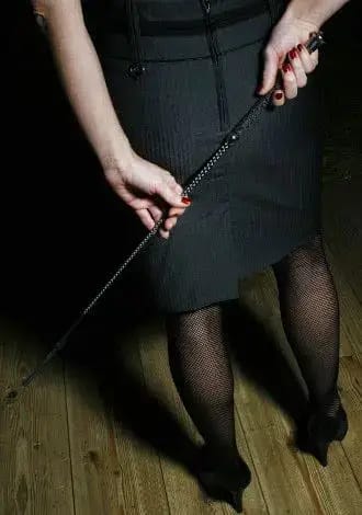 Mistress Danica hält eine schwarze Gerte vor ihrem schwarzen Kleid.