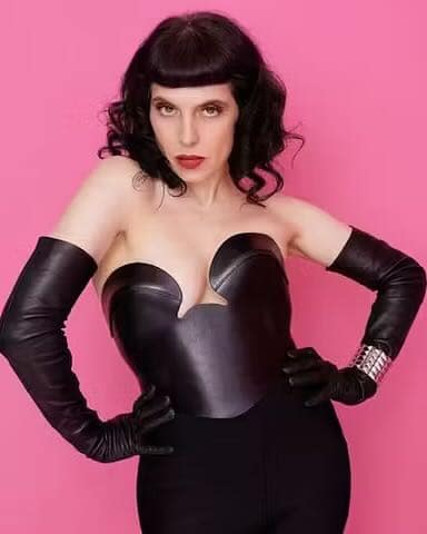 Portrait of Lady Vyra, a Berlin Dominatrix, wearing a black fetish outfit and long gloves against a pink background.