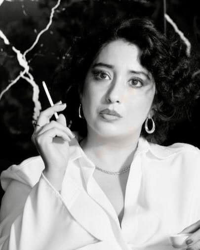 Black-and-white portrait of Mistress Leonor, a professional dominatrix in Barcelona, wearing a white blouse and hoop earrings, holding a cigarette and gazing thoughtfully to the side.