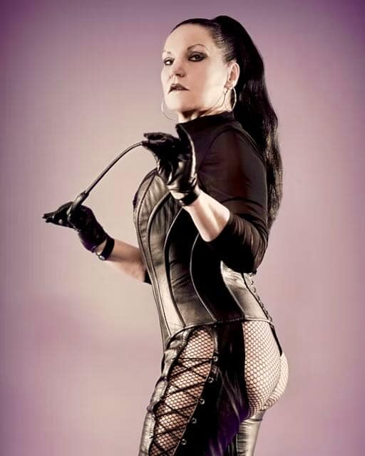 Mistress Silvia from Munich wearing a black leather outfit and fishnet stockings, holding a riding crop.