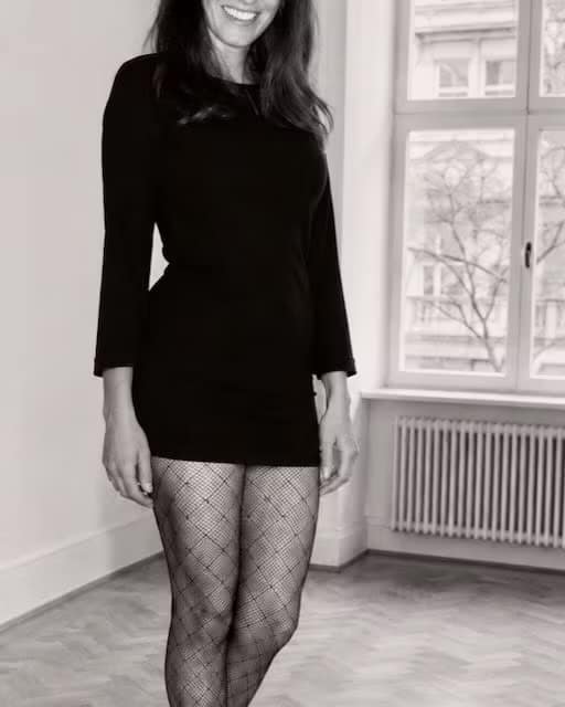 Findom Lady Judith from Berlin in a black dress, standing confidently by a large window.