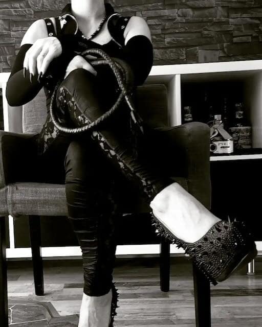 Goddess Helia von Roykamon, strict private FemDom, seated in black with a whip.