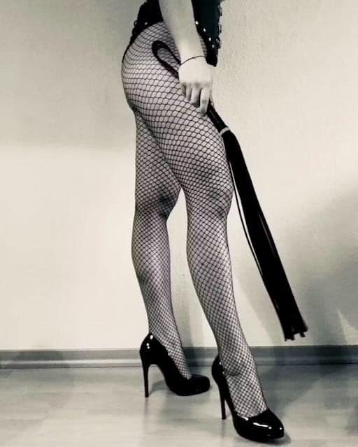 Lady Jay in fishnet tights and high heels holding a flogger