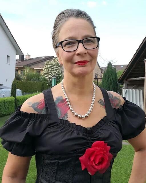 Private Femdom Lady Rose aus der Schweiz in schwarzem Outfit mit Brille und sichtbaren Tattoos.