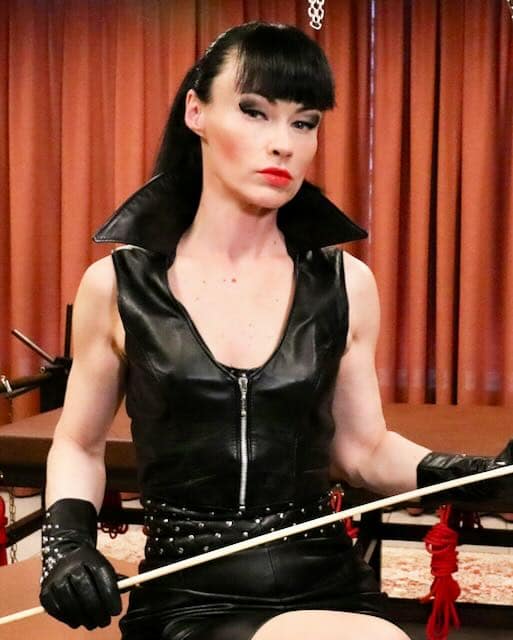 Cheyenne de Muriel in schwarzem Lederoutfit mit Handschuhen und Rohrstock.
