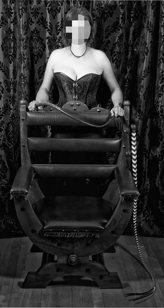Lady Sandra standing behind a dominant-style chair, holding a whip in a classic Femdom setting.
