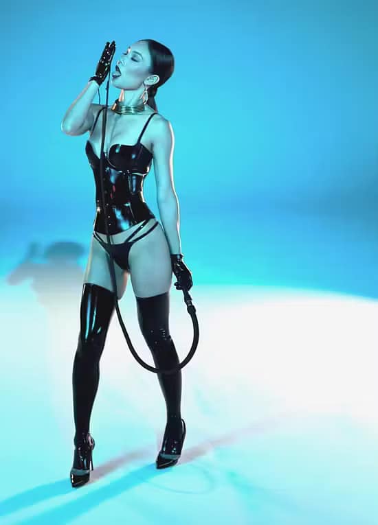 Mistress Gemma Li, Domina aus Colorado, USA, steht in schwarzem Latex-Outfit mit Overknee-Stiefeln, High Heels und Peitsche vor blauem Studiohintergrund.