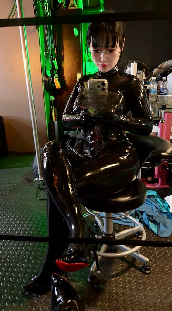 Mistress Hinako, Japanese dominatrix in black latex taking a mirror selfie in a BDSM studio