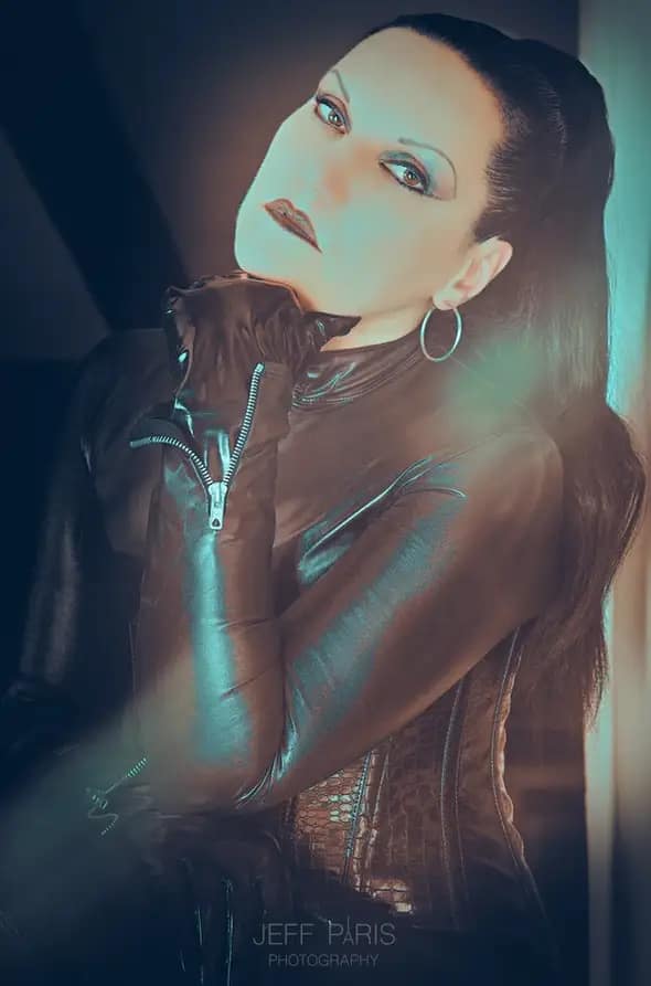 Portrait of Mistress Silvia in a black latex outfit and gloves, posing with an intense gaze and dramatic lighting.
