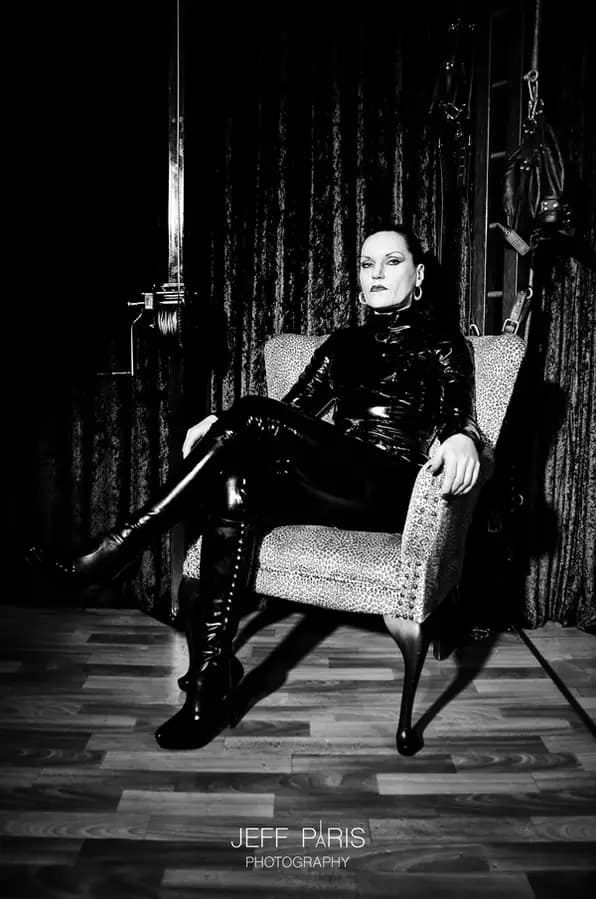 Mistress Silvia sitting confidently in a chair wearing a black latex outfit and high boots inside a dark BDSM studio.