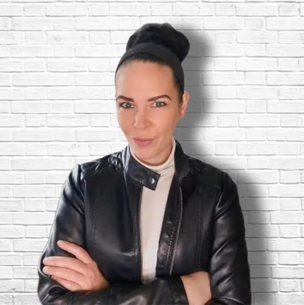 Portrait of BlackNightLady, a private femdom, standing with arms crossed in front of a white brick wall
