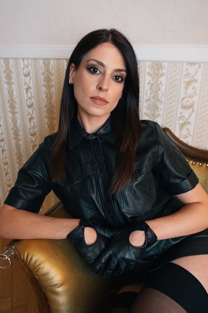 Mistress Aido Akida, a Berlin-based dominatrix, seated in a leather outfit and black gloves, looking directly into the camera.
