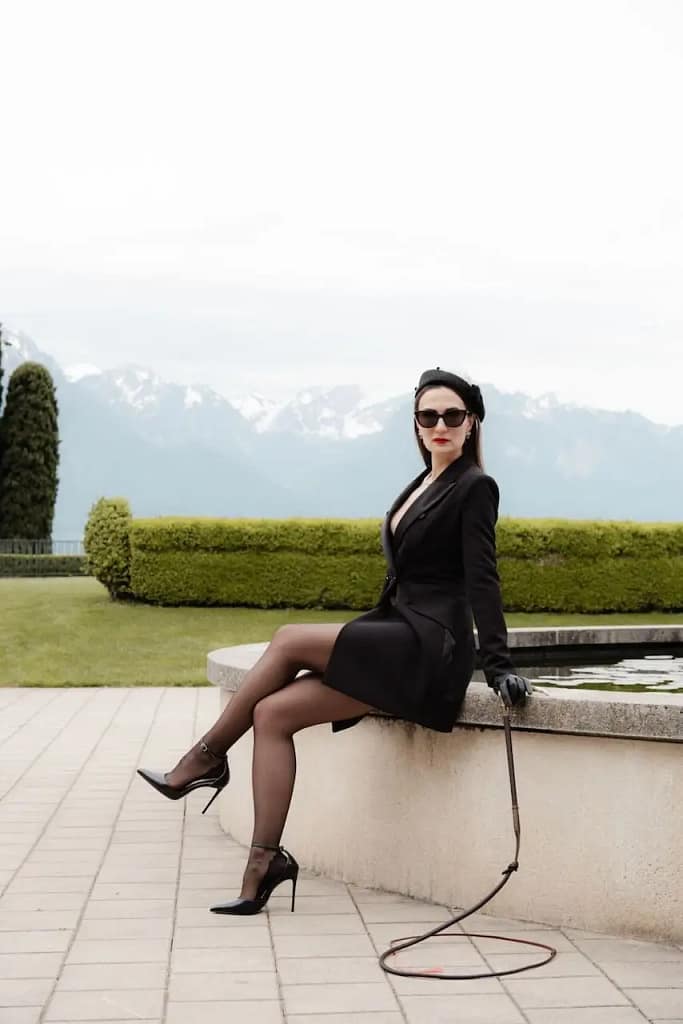 Domina Mistress Severna in schwarzem Mantelkleid, Sonnenbrille und High Heels mit Peitsche vor einer Bergkulisse bei Zürich.