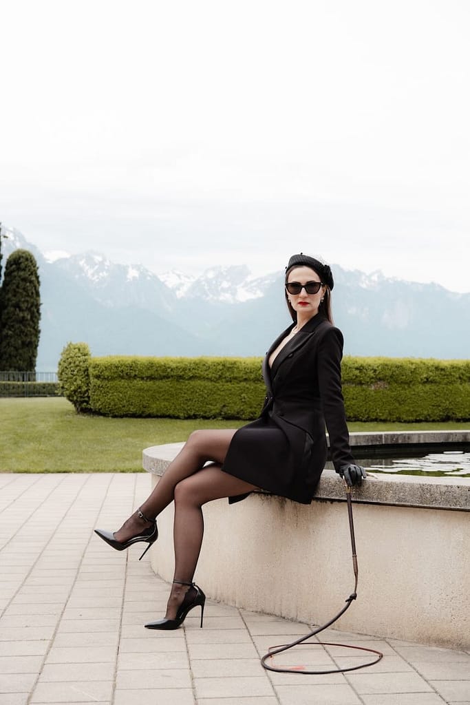 Mistress Severna, Dominatrix in Lausanne, Geneva, and Zurich, sitting outdoors by a reflecting pool with mountains in the background, wearing a black outfit, sunglasses, and high heels while holding a whip.