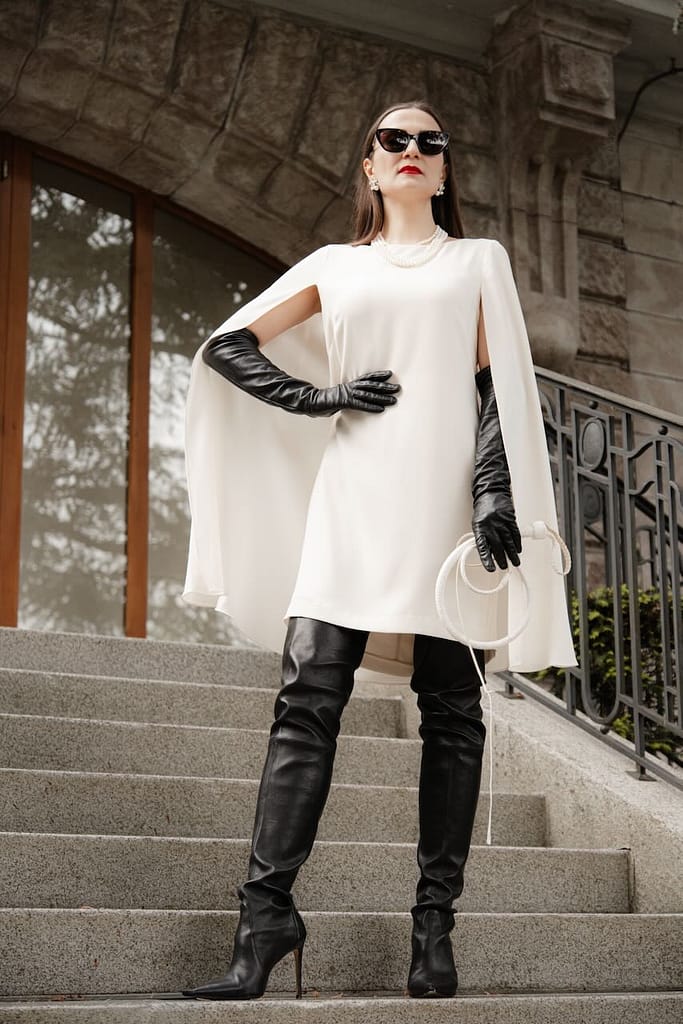 Mistress Severna, Dominatrix in Lausanne, Geneva, and Zurich, wearing a white cape dress with black gloves and thigh-high boots on a stone staircase.