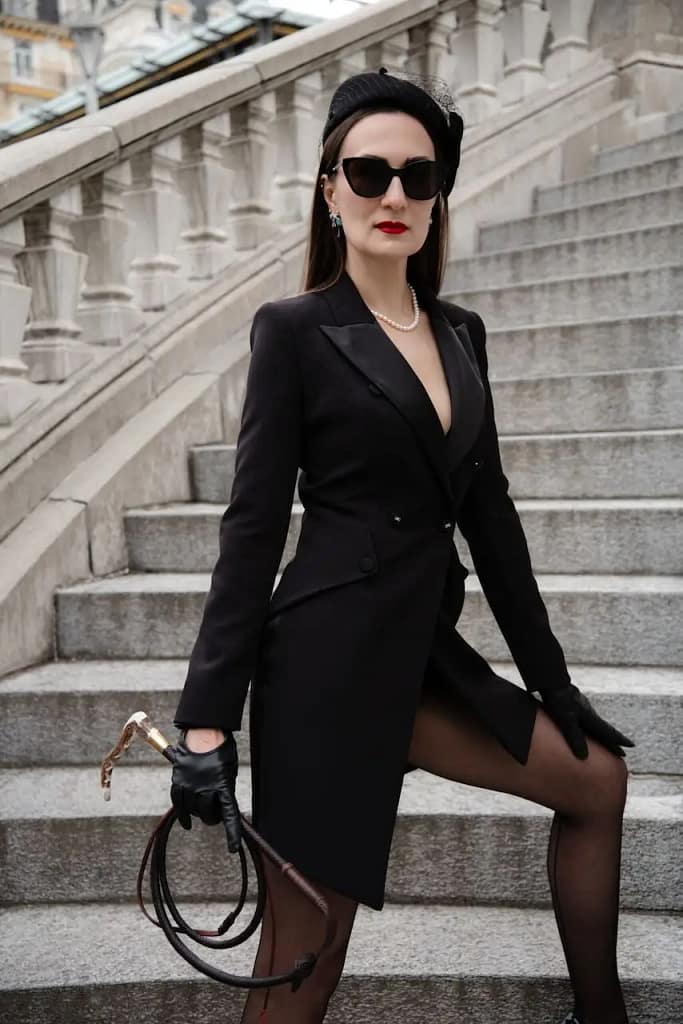 Domina Mistress Severna in schwarzem Mantelkleid, Sonnenbrille und Handschuhen mit Peitsche auf einer Steintreppe.