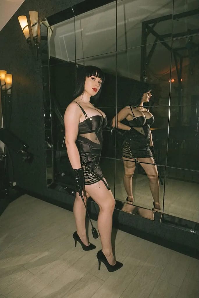 Mistress Felina, a dominatrix from Munich, posing in a black fetish dress and high heels inside a BDSM studio with mirrored walls.