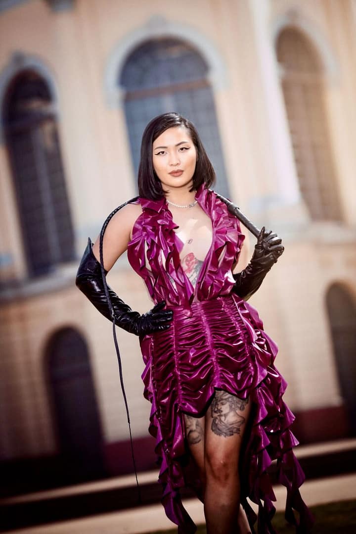 Kink Akita, Berlin Dominatrix, wearing a dramatic magenta latex dress and black gloves while holding a whip.
