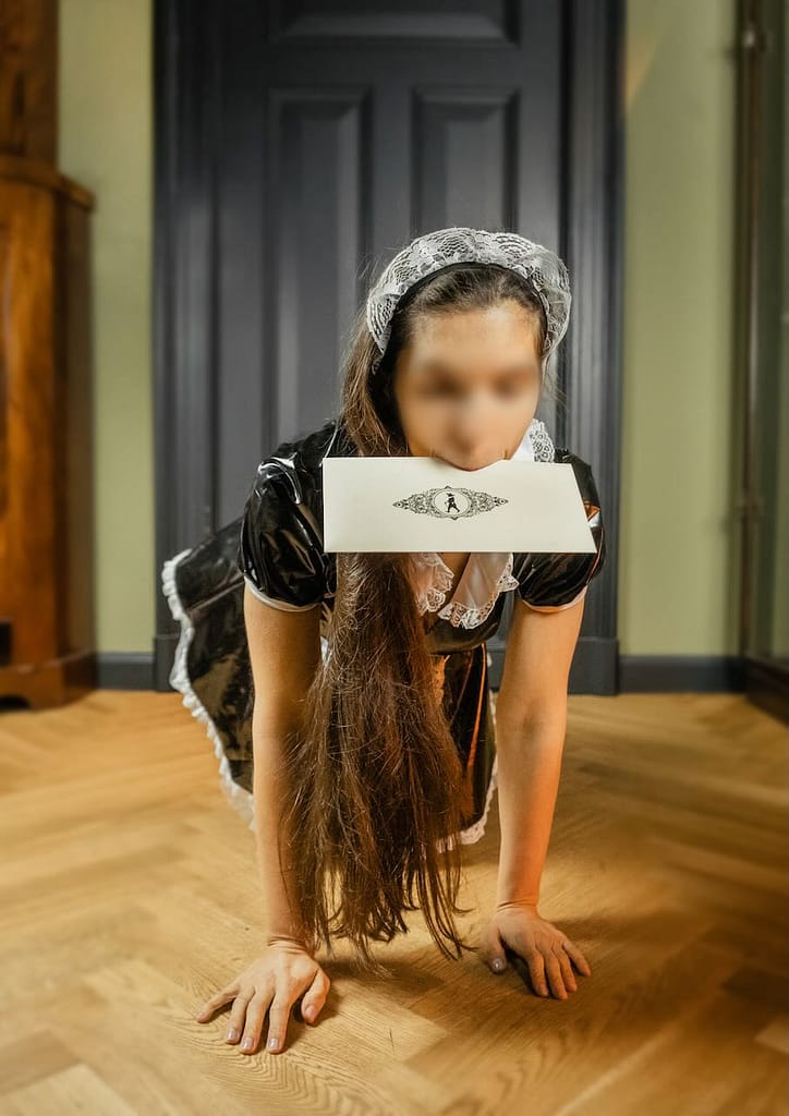 Submissive Fetish Escort Catherine Schneider kneeling on the floor in a maid outfit, holding a letter in her mouth, symbolizing submission and control