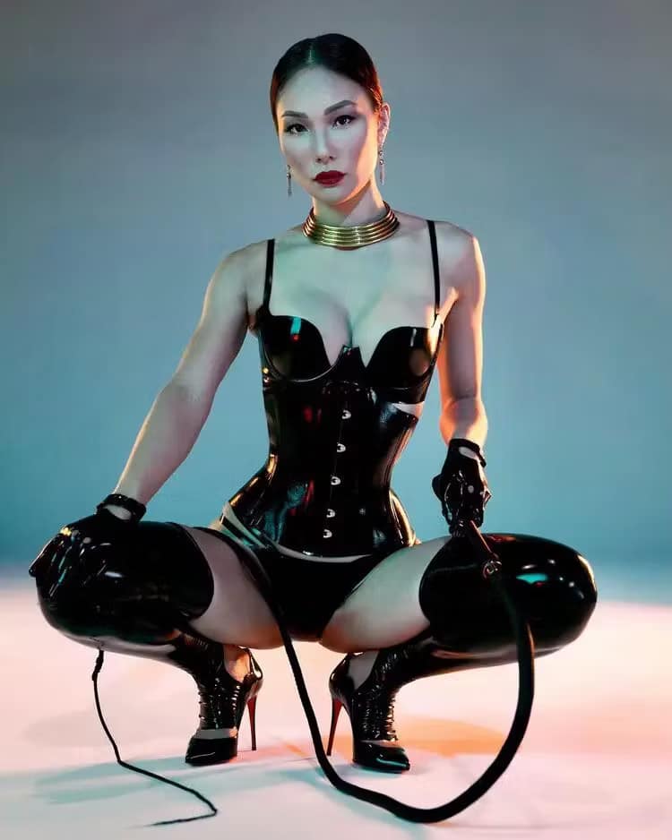 Domina in schwarzem Latex-Korsett, Latexhandschuhen und High Heels posiert selbstbewusst in der Hocke mit Peitsche.