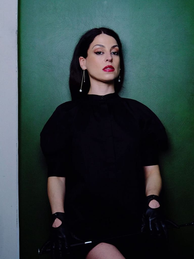 Mistress Aido Akida, a Berlin-based dominatrix, wearing a black dress, black gloves, and pearl earrings against a dark green wall.