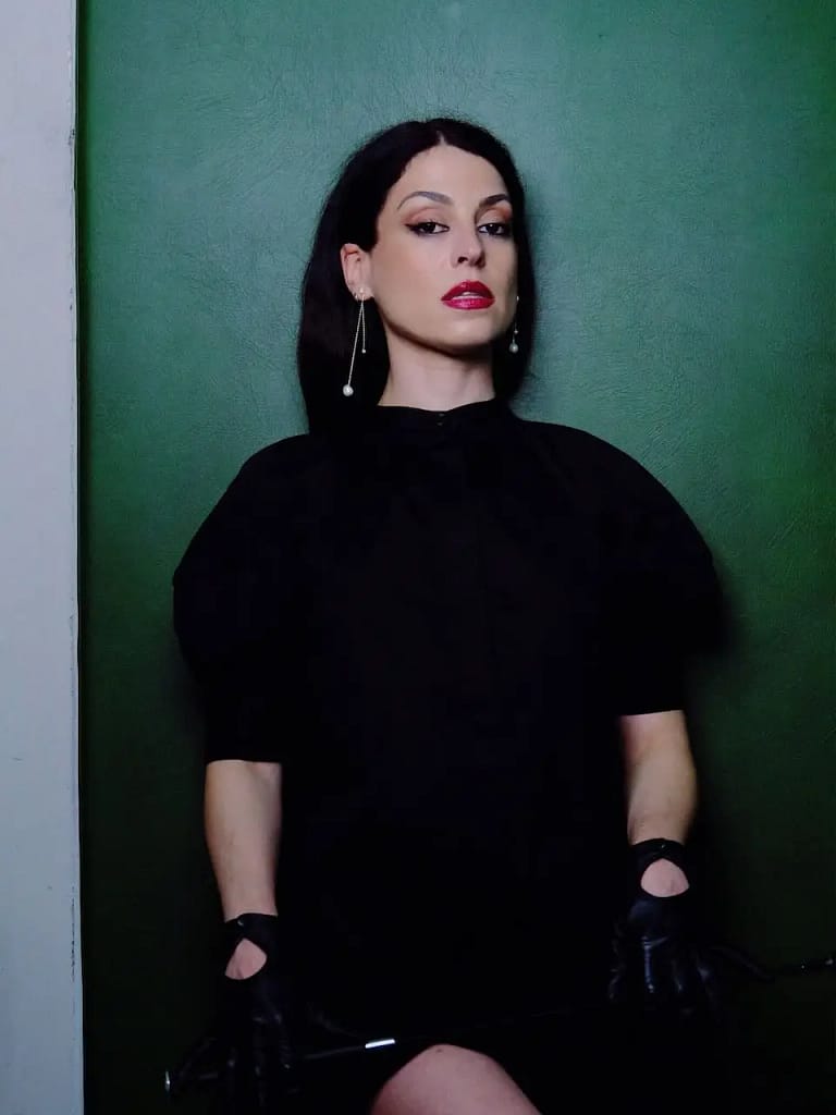 Mistress Aido Akida, Domina in Berlin, in schwarzem Outfit und Handschuhen vor einer grünen Wand.