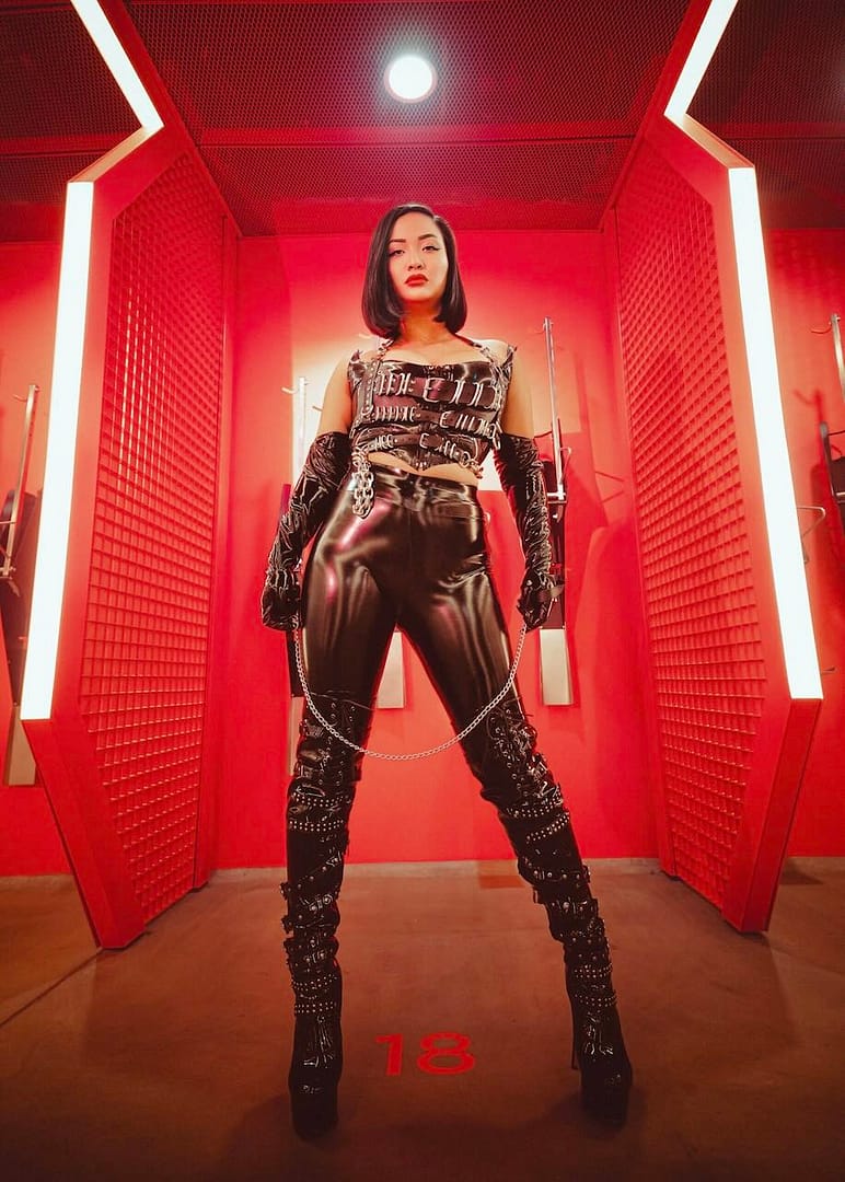 Full-length portrait of Kink Akita, a Berlin Dominatrix, wearing black latex and leather fetish attire in a dramatic red-lit BDSM setting.