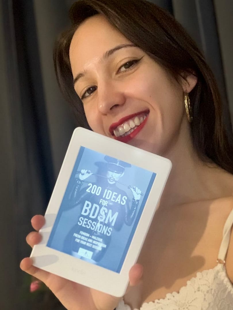 Mistress Fun from London, UK, smiling while holding an eBook reader displaying “200 Ideas for BDSM Sessions” by Lady Sas.