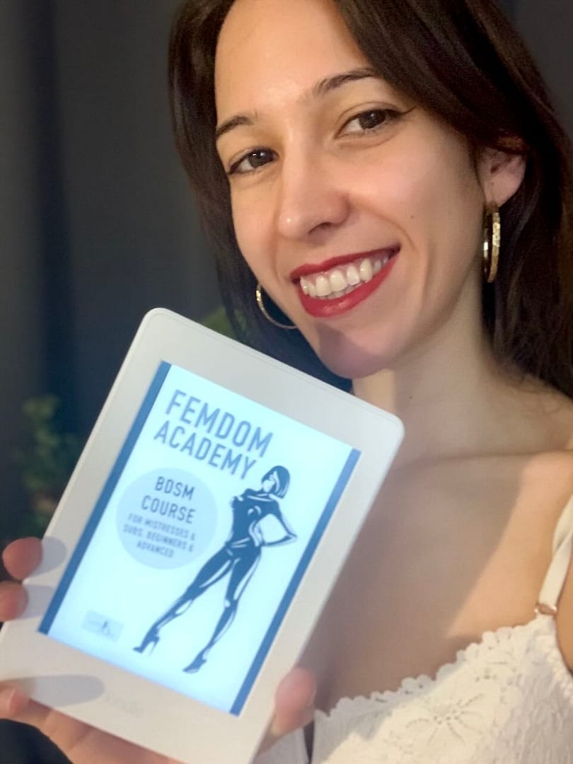 Mistress Fun from London, UK, smiling while holding an eBook reader displaying the book “Femdom Academy” by Lady Sas.