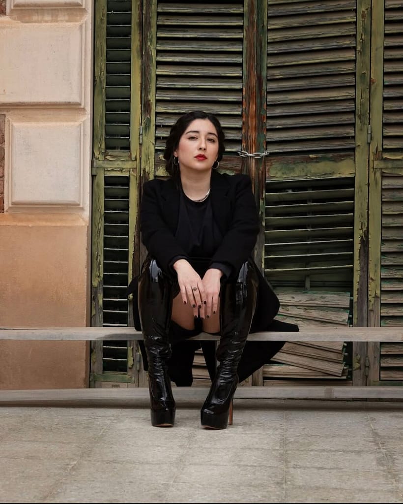 Mistress Leonor, a professional dominatrix in Barcelona, wearing a black blazer and glossy thigh-high platform boots, seated in front of weathered green wooden shutters.