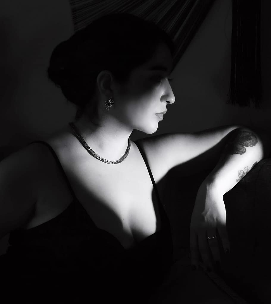 Black-and-white side-profile portrait of Mistress Leonor, a professional dominatrix in Barcelona, wearing a black spaghetti-strap dress and elegant jewelry, with a tattoo visible on her arm.