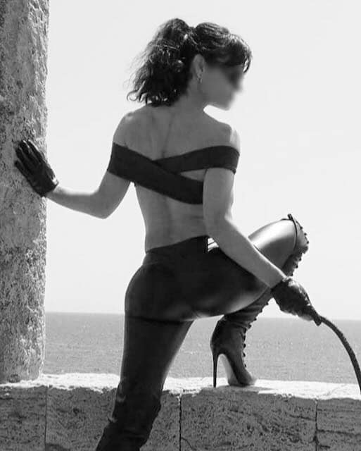 Domina Bess from Barcelona in a black-and-white portrait wearing thigh-high boots and holding a whip by a stone window overlooking the sea.